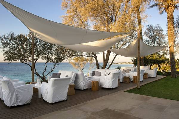 Außenterrasse des Grecian Sands Hotels mit weißen Möbeln und Sonnenschirmen am Meer