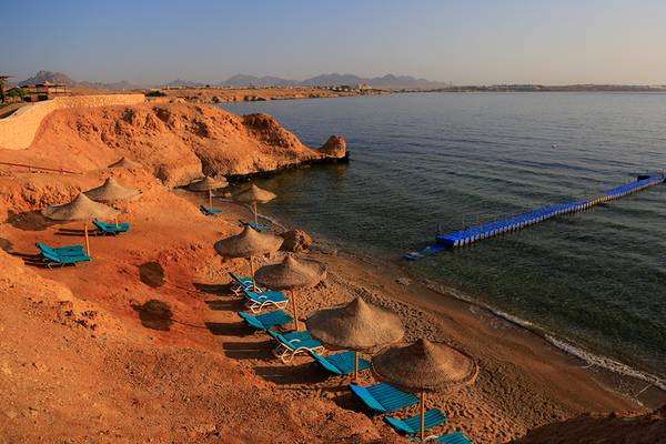 Strand des Sharm Club Beach Resorts mit Sonnenliegen, Sonnenschirmen und einer Brücke im Wasser.