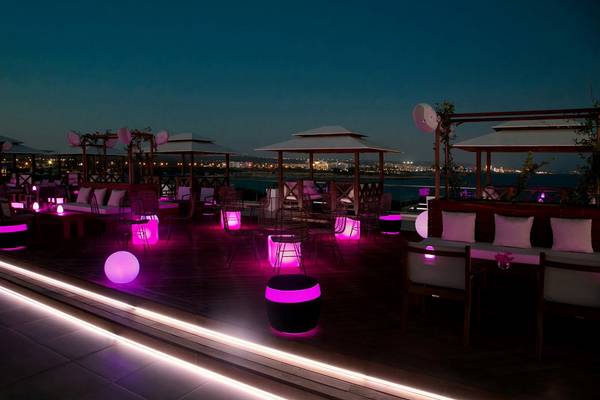 Abendliche Terrasse des Asterias Beach Hotels mit farbigen Beleuchtungselementen und Blick auf die Stadt.