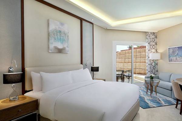 Innenansicht eines Zimmers im Hilton Hurghada Plaza Hotel mit einem großen Bett, einem Schreibtisch und einem Wohnzimmerbereich.