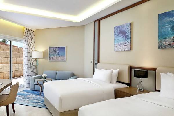 Innenansicht eines Doppelzimmers im Hilton Hurghada Plaza Hotel mit zwei Betten, einem Schreibtisch, einer Terrassentür und einer Sitzecke.