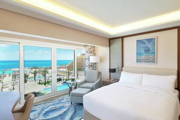 Zimmer im Hilton Hurghada Plaza mit Meerblick, modernes Interieur, große Fenster, Doppelbett, Nachttische, Sessel und Gemälde.