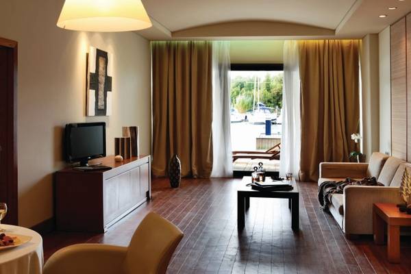 Wohnbereich des Porto Carras Meliton Hotels mit Fernseher, Tisch, Couch und offenem Fenster.