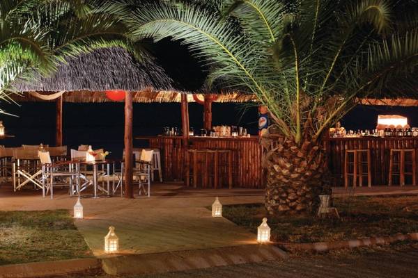 Nachtansicht der Terrasse des Porto Carras Meliton Hotels mit Bar, Tischen und Stühlen und Palmen.