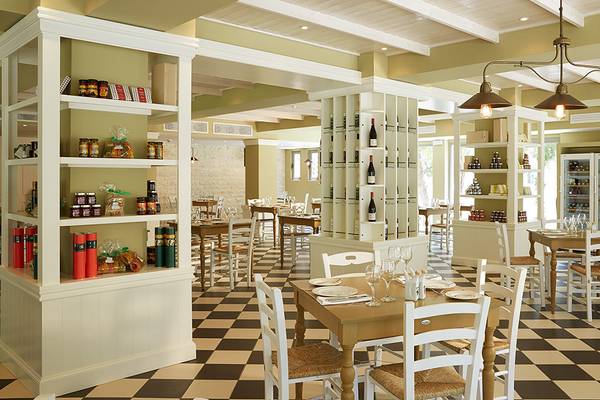 Innenansicht des MarBella Corfu Restaurants mit schachbrettartigem Boden, weißen Möbeln und Regalen mit Produkten.