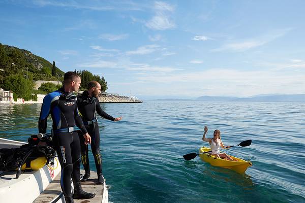 Zwei Taucher stehen am Dock, während eine Frau in einem gelben Kajak auf dem Wasser paddelt.