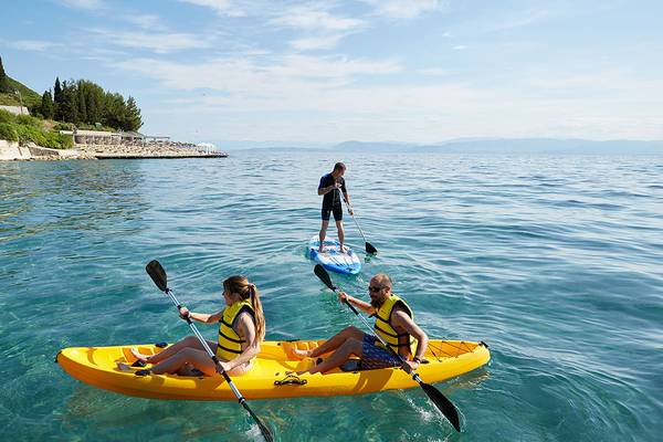 Ein Mann steht auf einem Surfbrett im Wasser, während zwei Paare in einem Kajak rudern, alles im Hintergrund des MarBella Corfu Hotels.