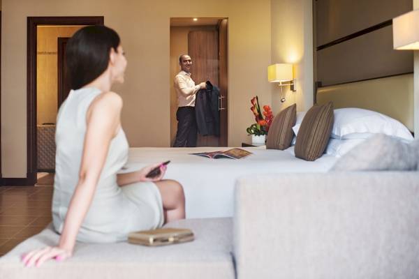 Eine Frau sitzt auf einem Sofa im Hotelzimmer, während ein Mann im Hintergrund ein Jackett anprobiert.
