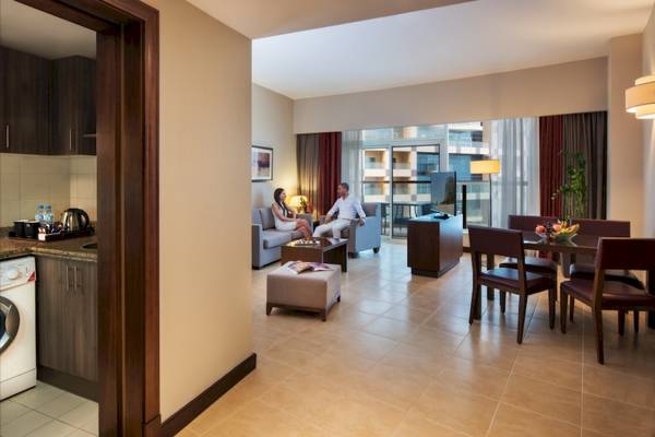 Zwei Personen sitzen auf einem Sofa im Wohnzimmer des Khalidiya Palace Rayhaan by Rotana, mit Blick auf einen Fernseher und ein Fenster.