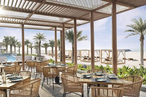 Außenterrasse des InterContinental Ras Al Khaimah Mina Al Arab Resort & Spa mit Tischen und Stühlen, überdacht von Holzsäulen und Palmen, Blick auf den Pool und den Strand.