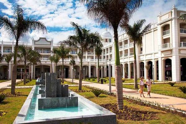 Außenansicht des Riu Palace Bavaro Hotels mit Brunnen, Palmen und zwei Personen, die vorbeigehen.