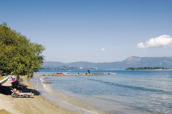 Strandszene am Louis Kerkyra Blue Hotel mit Liegestühlen, Bäumen und Bergen im Hintergrund.