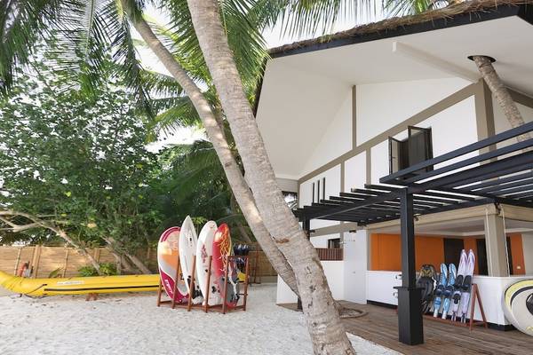Außenansicht des Dhawa Ihuru Hotels mit Wassersportgeräten und Palmen im Vordergrund.