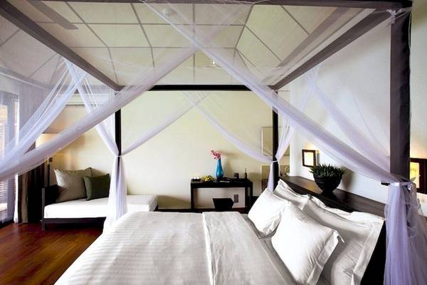 Schlafzimmer im Lily Beach Resort & Spa mit Himmelbett, weißen Laken und Kissen, einem kleinen Tisch mit Vase und Pflanzen.