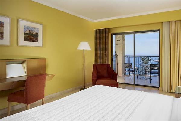 Ein modernes Hotelzimmer mit gelben Wänden, einem Bett, Schreibtisch, Stühlen und einer Terrasse mit Meerblick.