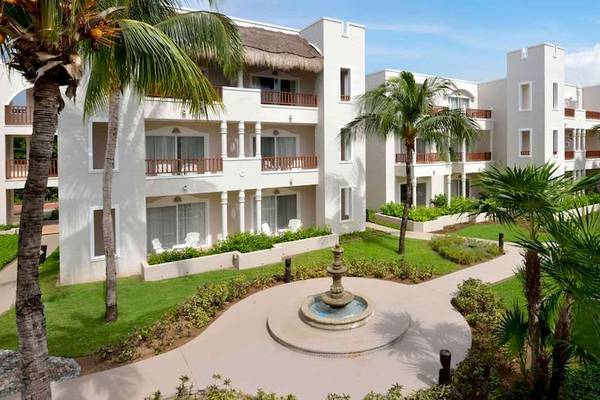 Außenansicht des weißen Catalonia Playa Maroma Hotels mit Palmen und einem Brunnen