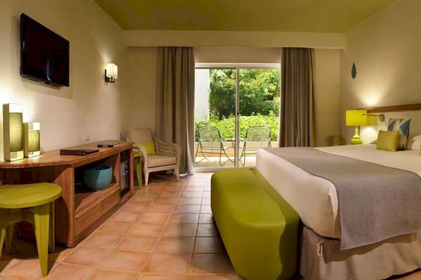 Hotelzimmer im Catalonia Playa Maroma mit Doppelbett, grünen Stühlen und Tisch, Fernseher und Balkonzugang.