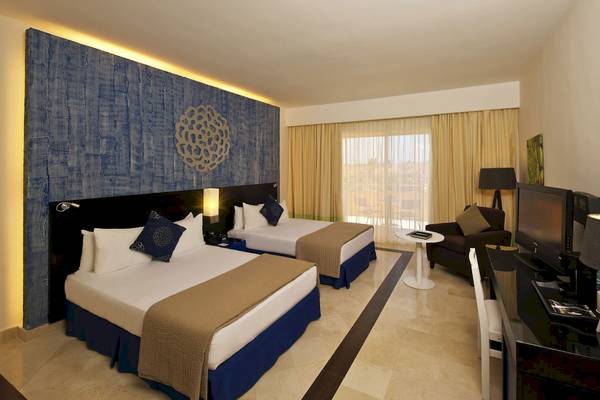 Doppelzimmer im Ocean Maya Royale mit zwei Betten, Nachttischen, Lampen, Fernseher und einem Fenster mit Vorhängen.