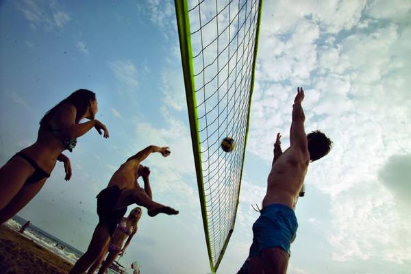 Mehrere Personen spielen Volleyball auf einem Strand, einer Spieler springt um das Netz.