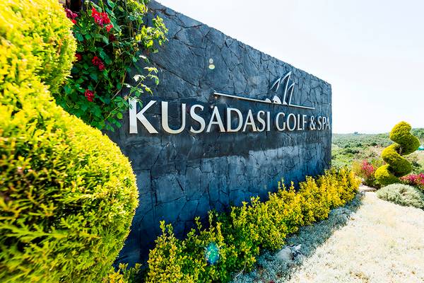 Eingangsschild des Kusadasi Golf & Spa, umgeben von grünen Büschen und Pflanzen.