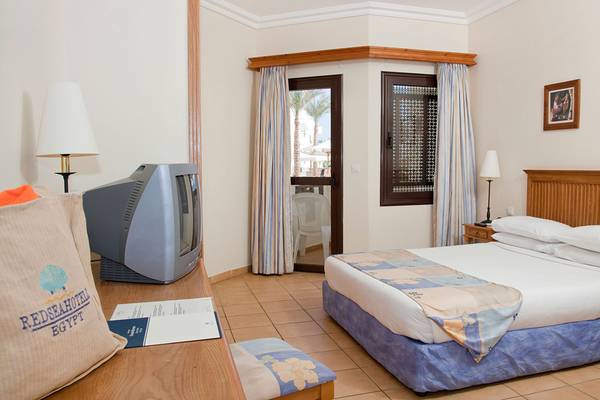 Ein Hotelzimmer im Sharm Resort mit einem Bett, Fernseher, und einem Fenster mit Aussicht auf Palmen.