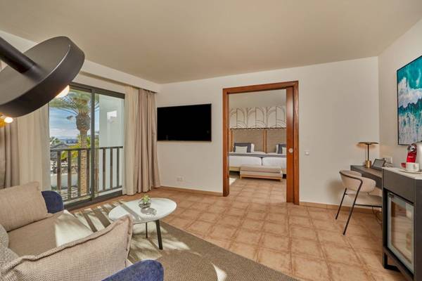 Innenansicht der Suite im Secrets Lanzarote Resort & Spa mit Wohnzimmer, Couch, Tisch und Balkon.