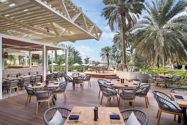 Außenterrasse des Le Meridien Mina Seyahi Beach Resort & Waterpark mit Tischen und Stühlen, umgeben von Palmen und Blick auf das Meer.