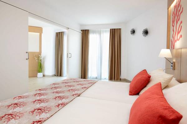 Schlafzimmer im Perla Hotel mit zwei Betten, roten Kissen und weißem Laken, einem Tischlampen und einer Wand mit Kunstwerk.