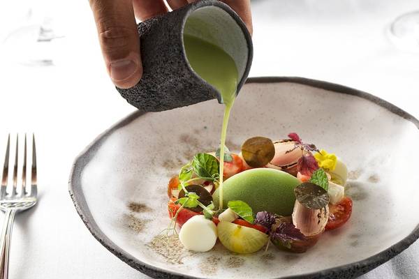 Ein Tellergericht wird mit grüner Fl üssigkeit aus einem Becher übergossen, serviert auf einem weißen Tisch.