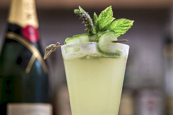 Ein Glas mit einem Cocktail, garniert mit Gurkenscheiben und Minze, vor einer Flasche Champagner.