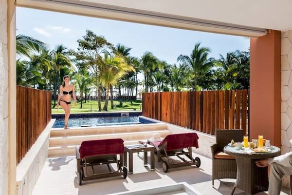 Ein Mann liest auf der Terrasse des Barcelo Maya Beach Hotels, neben einem Pool mit einer Frau in einem Bikini.