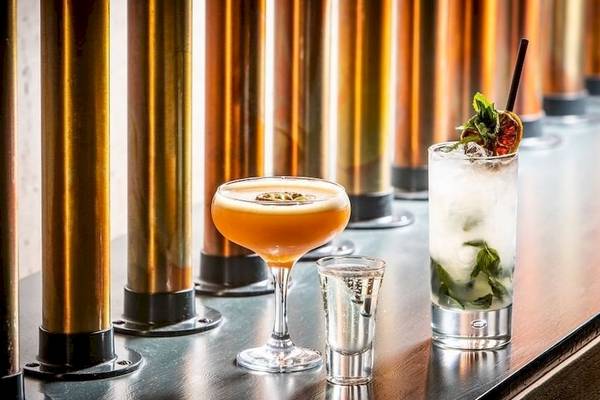 Drei Cocktails auf einer Bar mit goldenen Säulen, darunter ein Orangen-Cocktail, ein Mojito und ein Shot.