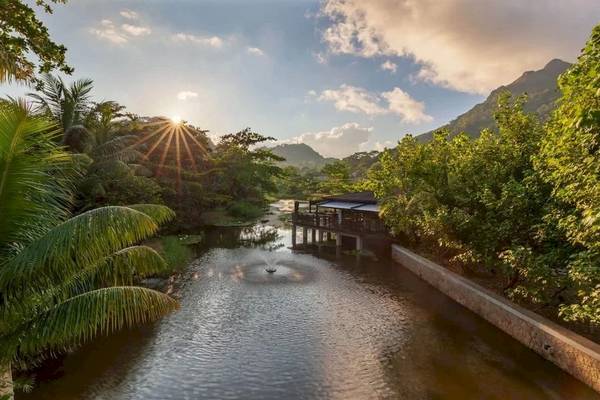Tropischer Fluss im STORY Seychelles Hotel bei Sonnenaufgang mit Gebäuden und Palmen im Hintergrund