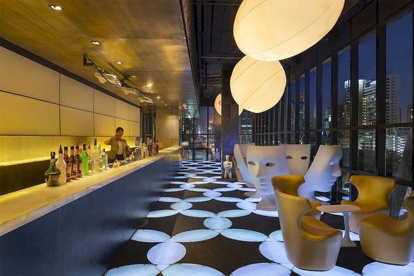 Innenansicht der Bar im Mode Sathorn Hotel mit Theke, Flaschen, goldenen Stühlen und blau beleuchteten Gesichtsmasken auf dem Boden.