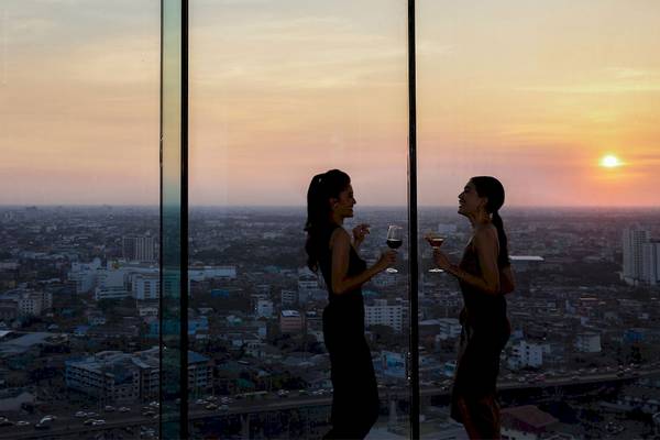 Zwei Frauen in Abendkleidern trinken Wein und genießen den Panoramablick auf Bangkok vom AVANI+ Riverside Bangkok.