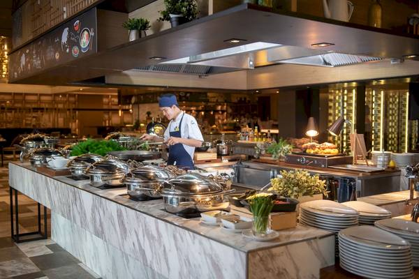 Ein Koch bereitet ein Buffet im Novotel Bangkok Sukhumvit 20 zu, mit Silberschüsseln und Gerichten auf dem Tisch.
