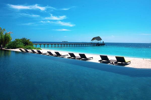 Ein großer Pool am Strand des Amari Havodda Maldives mit Sonnenliegen und einer Pier im Hintergrund.
