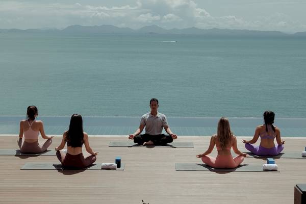 Ein Mann und vier Frauen machen Yoga auf einem Holzdeck am Strand mit Blick auf das Meer.