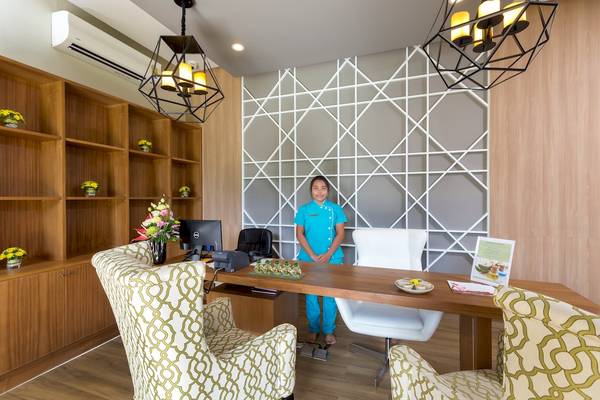Rezeption des X10 Khaolak Resorts mit einer Frau in blauer Uniform, die lächelt und einen Schreibtisch mit Blumen und Büchern hinter sich hat.
