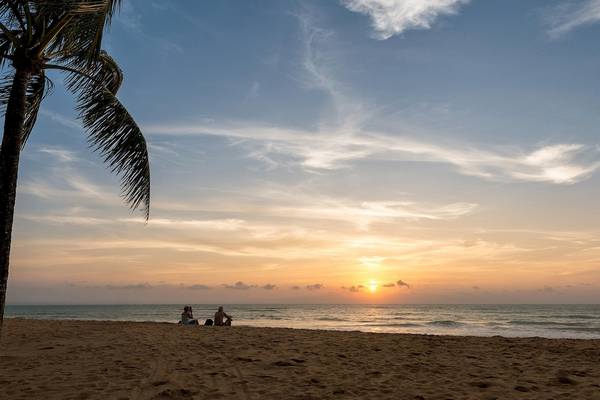 Zwei Personen sitzen am Strand und beobachten den Sonnenuntergang mit einer Palme im Vordergrund.