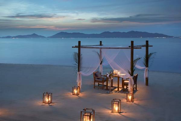 Ein romantisches Abendessen am Strand im Santiburi Koh Samui mit einem Tisch und Stühlen unter einer Pergola und Laternen im Sand.