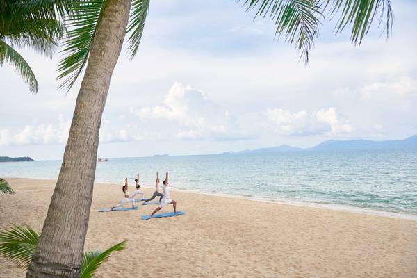 Vier Personen machen Yoga auf einem Strand vor dem Santiburi Koh Samui Hotel.