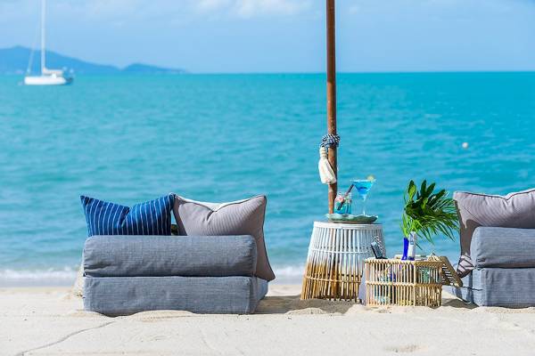 Ein Strand mit blauen Sofas, Kissen, einem Sonnenschirm und einem Tisch vor einem Meer und einer Segelyacht im Hintergrund.