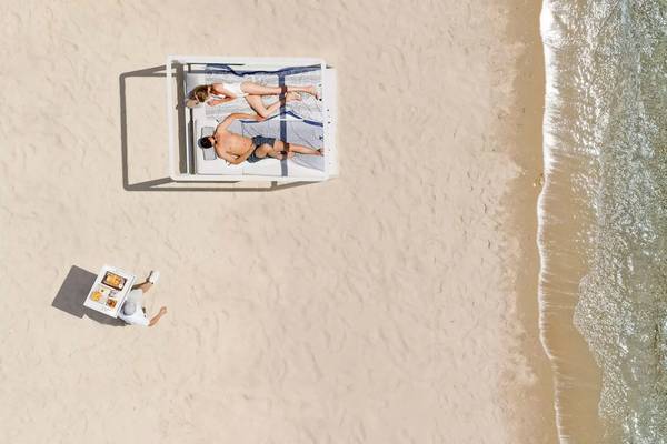 Zwei Personen liegen auf einem Bett am Strand mit einem Servicemitarbeiter, der ein Tablet hält.