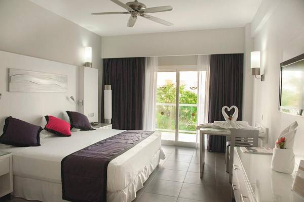 Innenansicht eines Hotelzimmers im Riu Palace Costa Rica mit einem großen Bett, Tisch und Glastür.