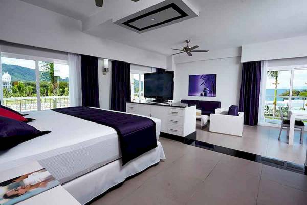 Innenansicht einer Suite im Riu Palace Costa Rica mit großem Bett, Fernseher und Blick auf den Garten.