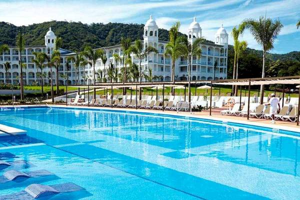 Ein großer Swimmingpool vor dem Riu Palace Costa Rica Hotel mit weißen Sonnenliegen und Palmen.