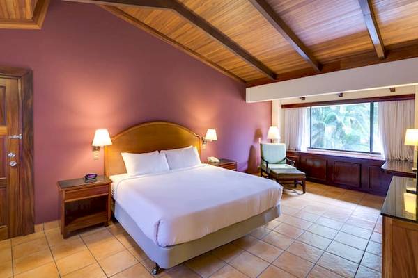 Doppelzimmer im DoubleTree by Hilton Hotel Cariari San Jose mit Bett, Nachttischen, Lampe, Fenster und Holzmöbeln.
