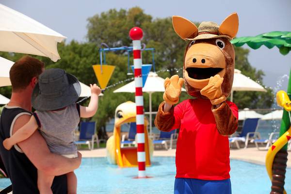 Ein Vater hält ein Kind im Arm am Schwimmbadrand, während ein Maskottchen ihnen zuwinkt.