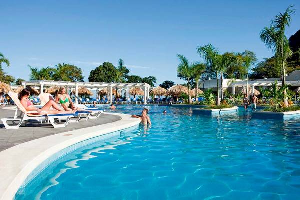 Hotelpoolszene mit Gästen, die auf Sonnenliegen entspannen und andere Gäste im Wasser sind.